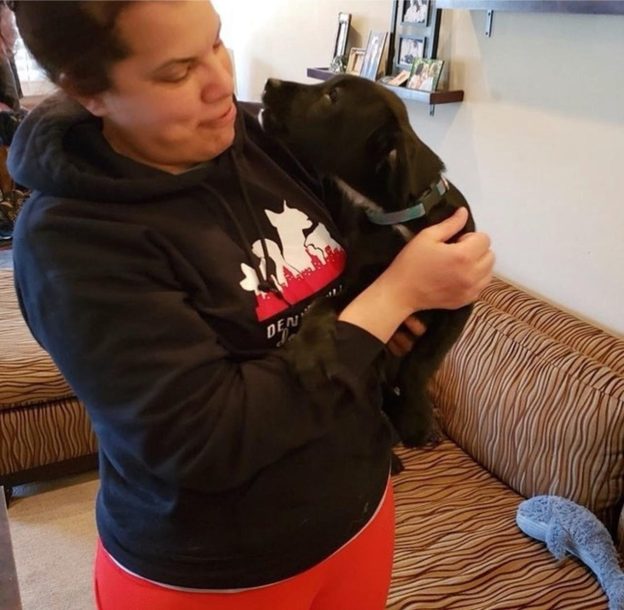 Denise Willis in a DW Dog Training hoodie, holding a black Labrador up against her shoulder in a living room and looking at him.